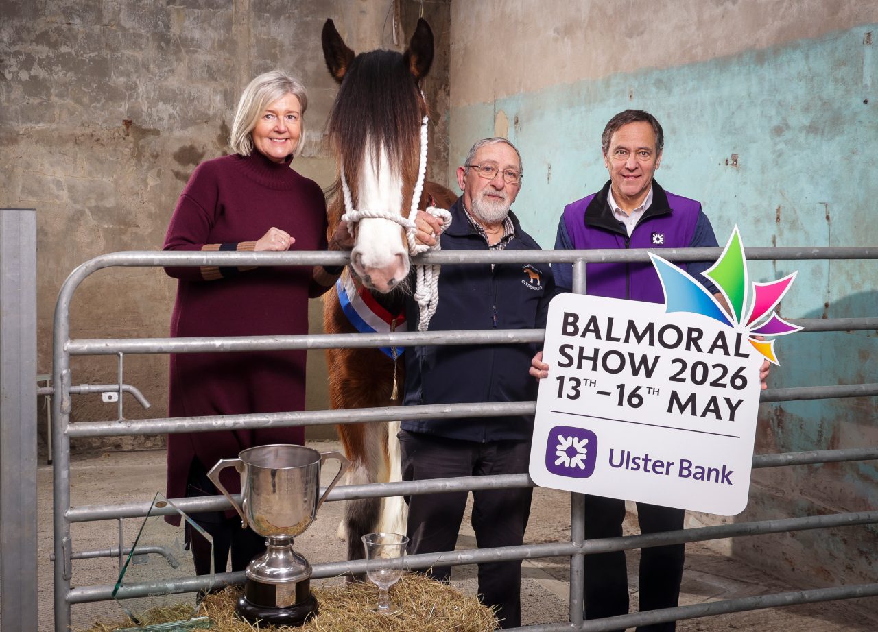 This image shows 2 men, a woman and a hourse in a stable at the Balmoral show 2026 with a metal winners cup, a glass trophy and a empty glass.