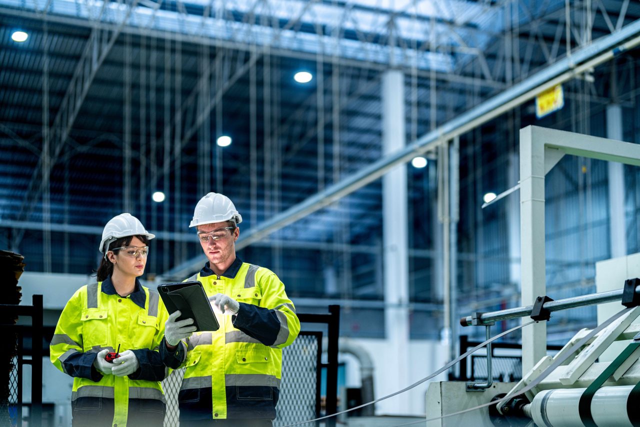 This image shows two people wearing hard hats and high visibility jackets in an industrial setting and looking at a tablet device. 