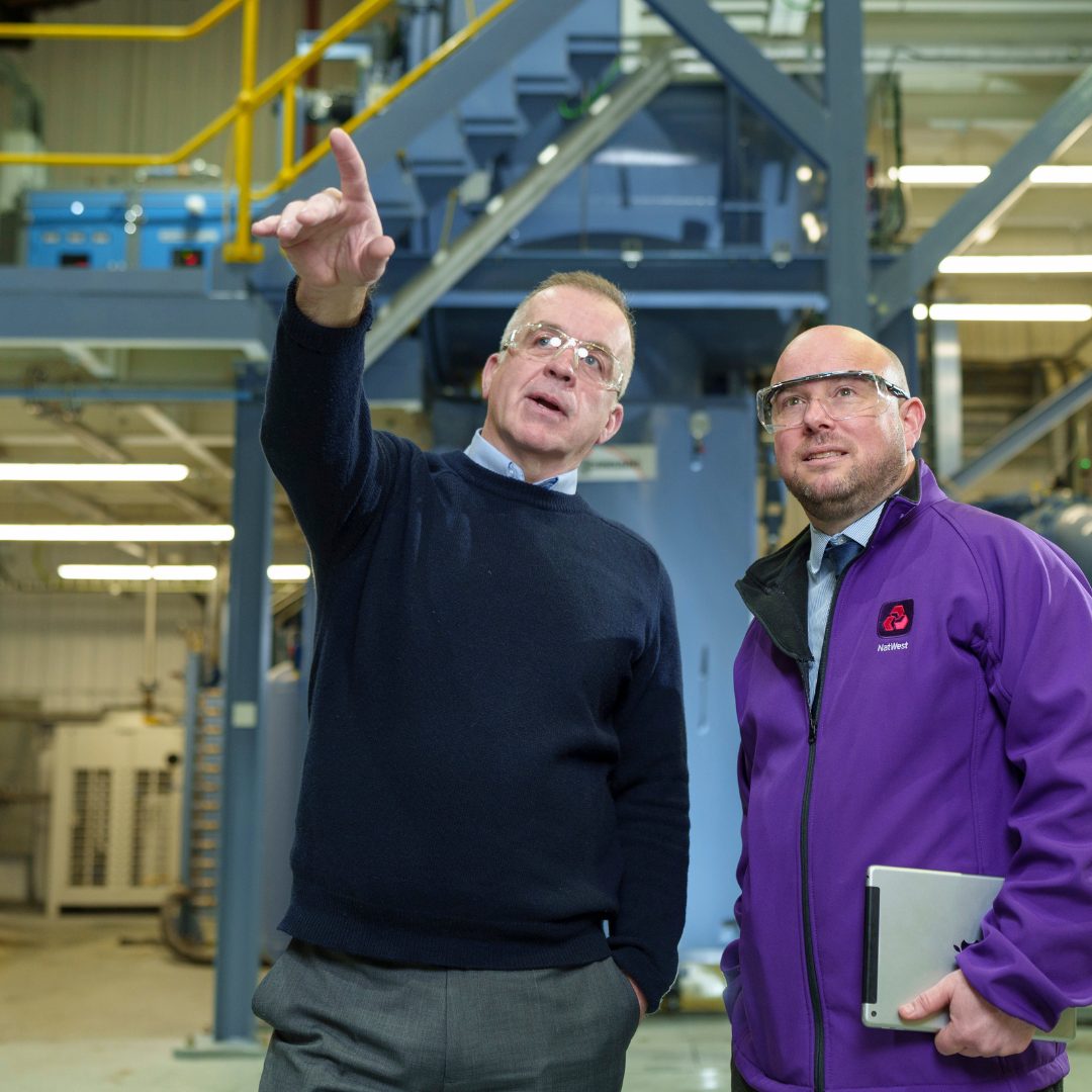 This image shows two men standing in a factory. They are pointing into the distance.