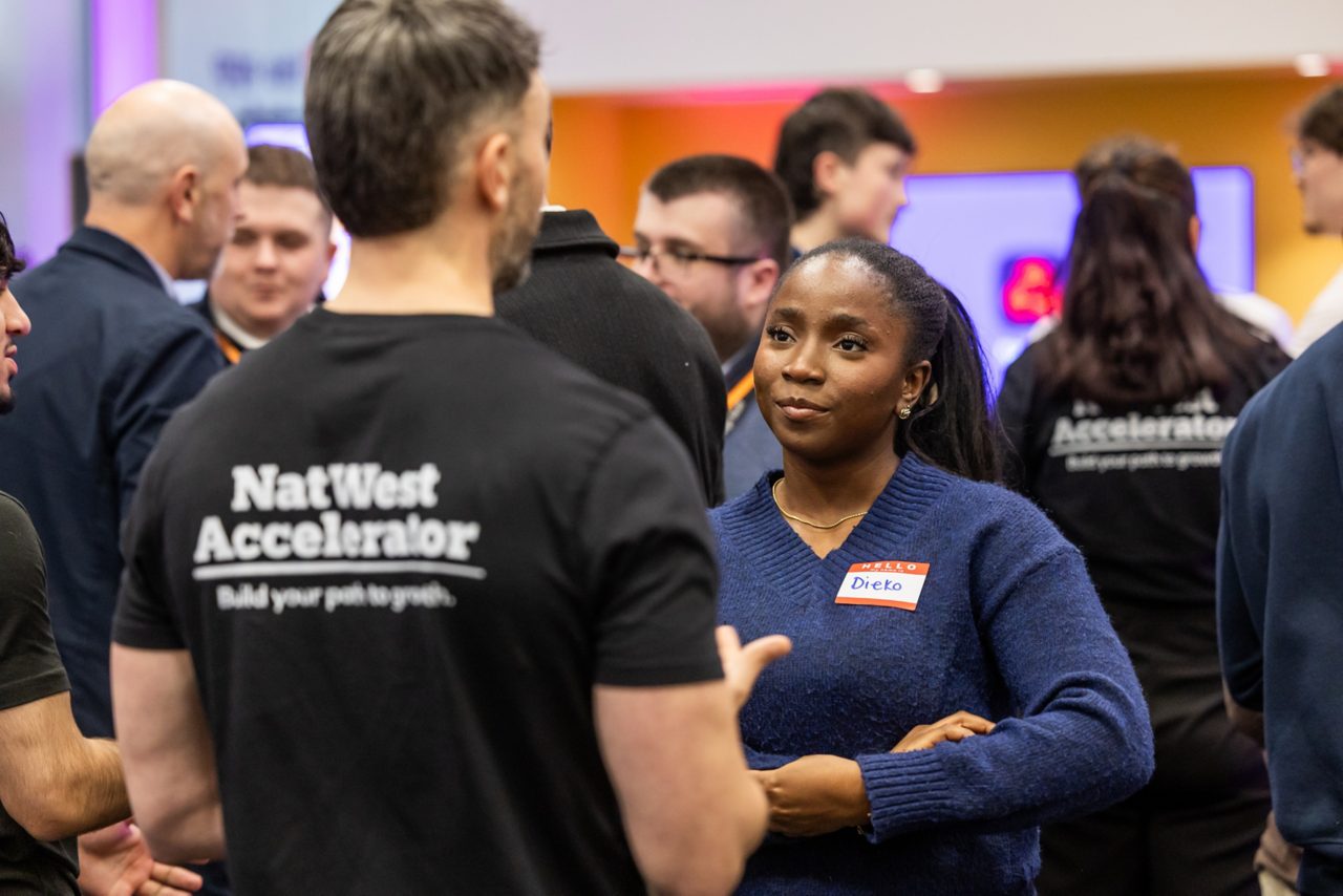 Image of people standing in a NatWest Accelerator. A man has his back to the picture while he faces a woman. The back of his t-shirt says, ‘NatWest Accelerator’.