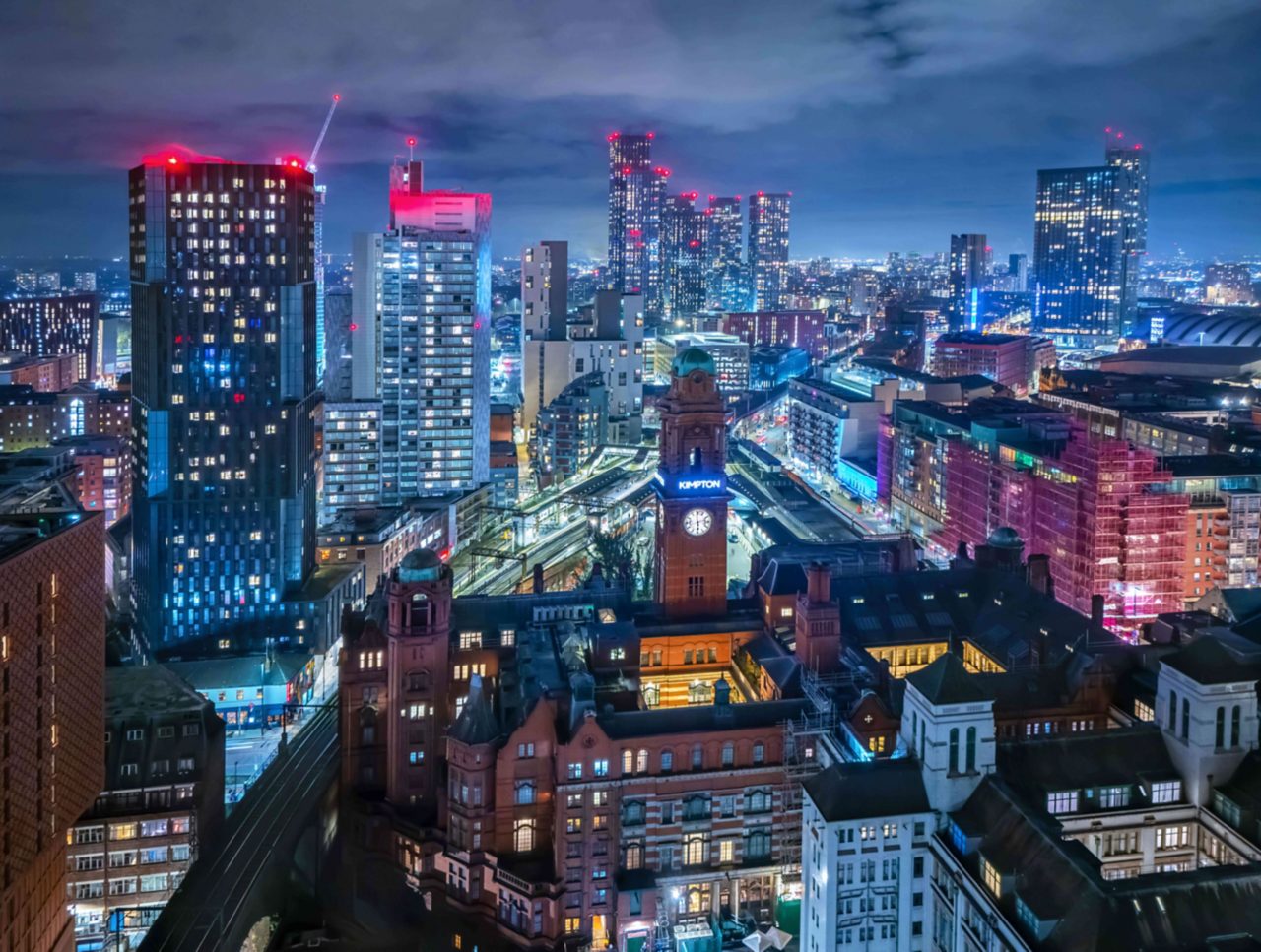 Nighttime cityscape of Manchester featuring the illuminated Central Library clock tower, surrounded by modern high-rises and vibrant city lights