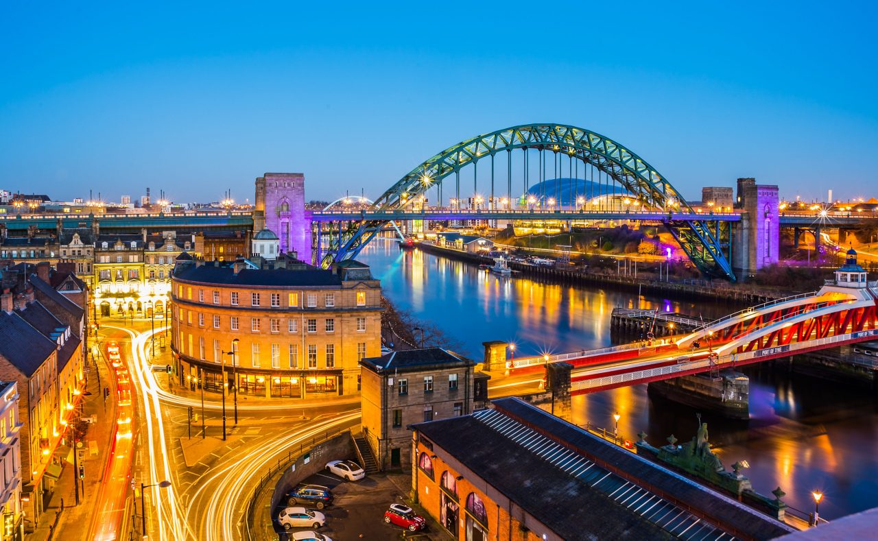 Evening view of Newcastle upon Tyne featuring the illuminated Tyne Bridge and Swing Bridge over the River Tyne, with city lights and vehicle trails