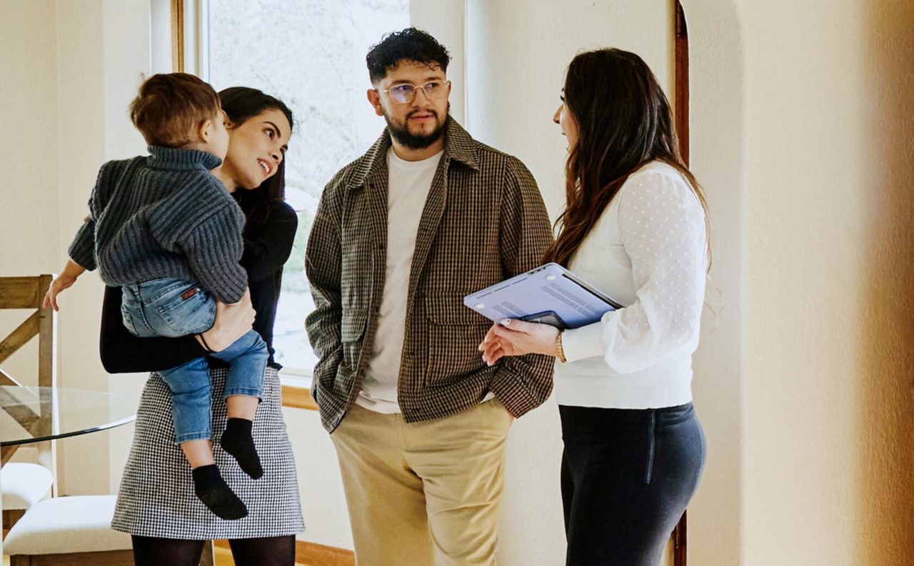 this image shows a man and a woman talking to another woman in a house. One of the woman is holding a baby. 