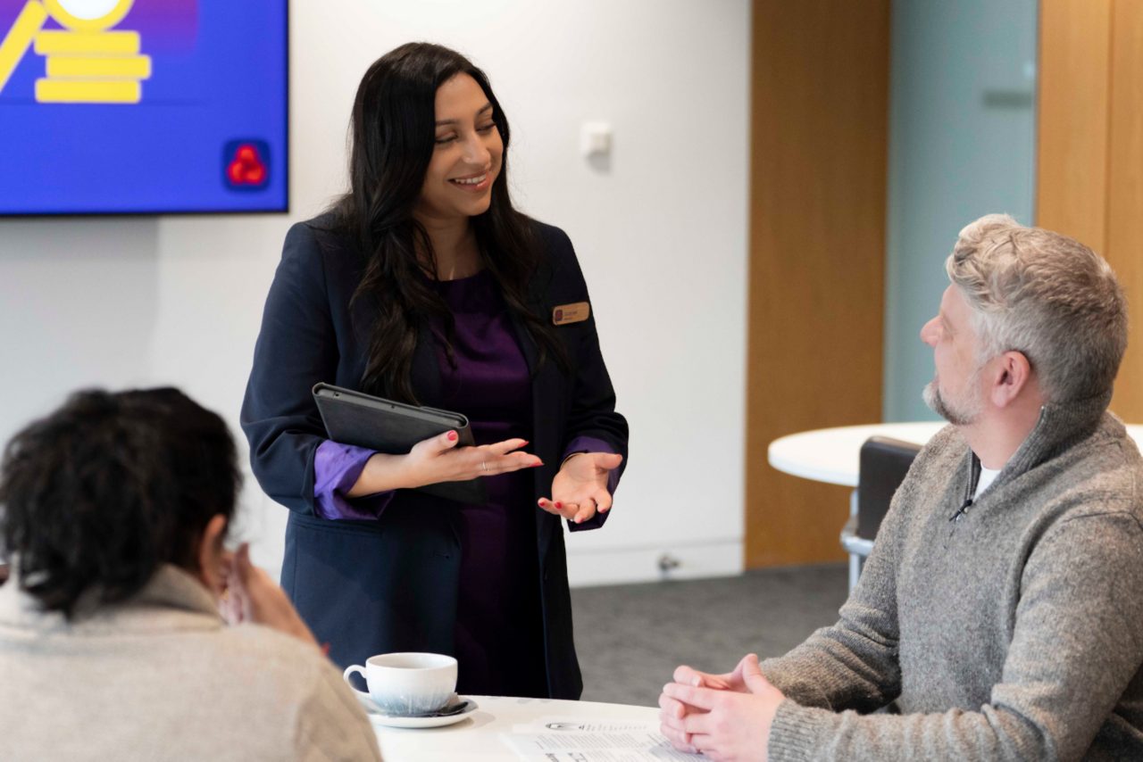 A person in business attire stands holding a tablet and speaking to two seated individuals at a table. A presentation screen with an icon is visible in the background, and a cup and documents sit on the table in front of the group.