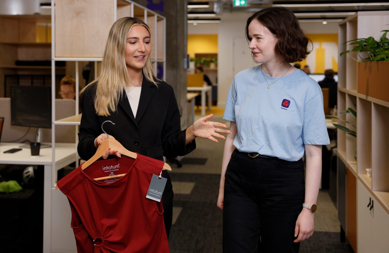 Two people are standing and talking in a modern office space. One person is holding a red dress on a hanger with product tags visible, while the other listens. Shelving units, desks, and office lighting appear in the background.